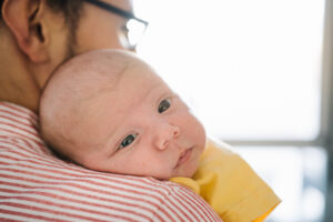 A close up of a baby’s face resting on a man’s shoulder. The baby is white skinned and wears a yellow t shirt. We see the back of the man’s shoulder and part of his face, he wears glasses and a red and white striped shirt