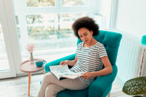 A mixed race woman sits in a comfy green armchair and reads a magazine