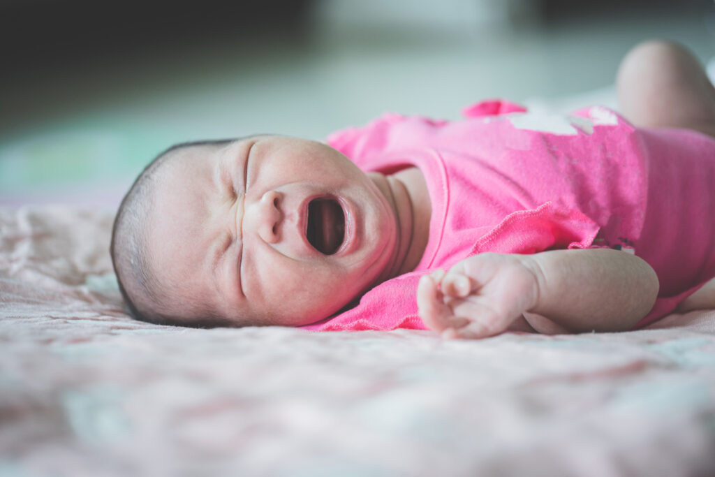 A baby in a pink onesie lies on a soft focus background. The baby is lying on its back with eyes screwed up and mouth wide open, crying.