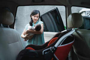 A young East Asian woman cradles her newborn as she stand by an open car door. She prepares to place the baby into the red and black car seat in the foreground of the photo.