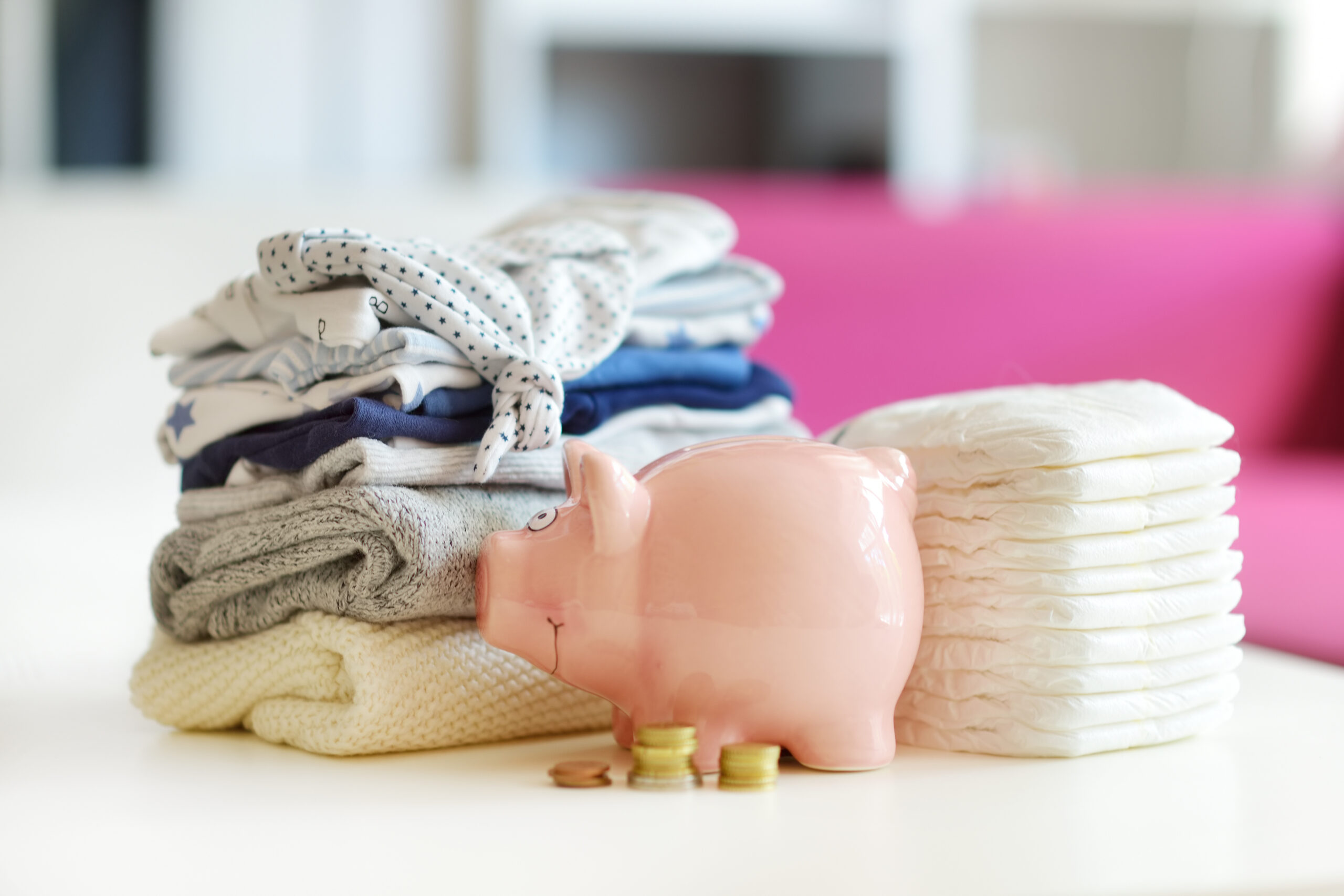 A pile of folded baby clothes in blue, grey and white sits next to a pile of disposable nappies. In front of this is a pink piggy bank with three small piles of coins in the foreground.