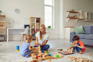 A blond haired white skinned woman sits on the floor playing with two young children. They play with a wooden train set and building blocks.