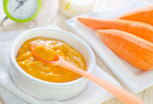 A white bowl of carrot purée with an orange fork and a white plate of carrots sit side by side in front of an out of focus white and green clock.
