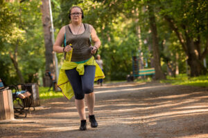 A white lady in leggings and a vest top with a bright yellow jacket tied round her waist is jogging along a path lined with trees. She carries a bottle of water in one hand and her phone in the other which is connected to earphones.