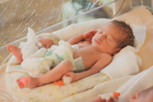 A tiny, white skinned baby with dark hair lies in an incubator wearing just a nappy. The baby has an IV in its hand and a foot monitor on.
