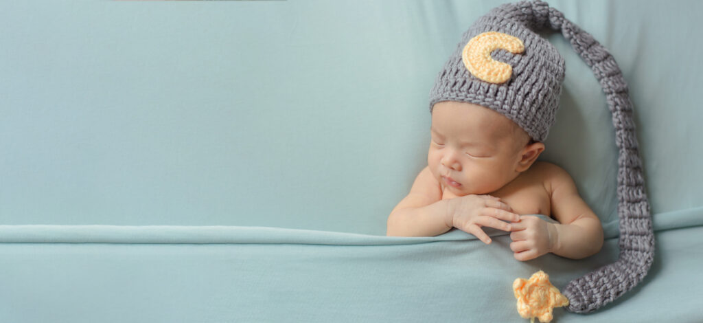 A sleeping baby in a woolly hat lies between muted green sheets