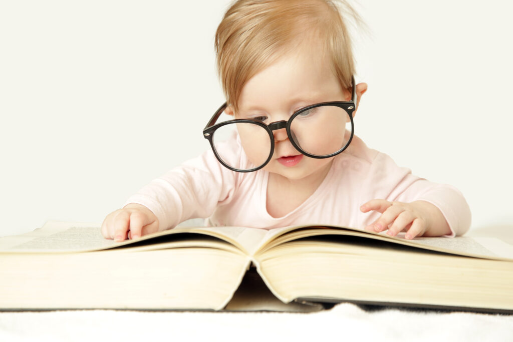 A blond baby is looking at a very large thick book. The baby is wearing oversized black rimmed glasses
