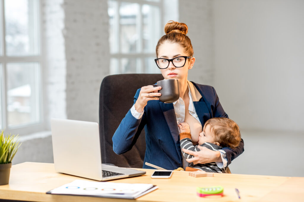 A mum is sitting at a desk drinking coffee and breastfeeding a baby during her break